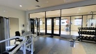 This single-floor fitness center is thoughtfully designed to accommodate a variety of workout styles. The space is divided into distinct zones separated by large, floor-to-ceiling glass panel partitions with sleek black framing, allowing for visual openness while maintaining functional separation between areas. The main workout area features rubberized flooring and houses strength training equipment, including a multi-function cable machine and resistance-based exercise stations. Through the glass partitions, a dedicated studio or stretching room is visible, equipped with stability balls, a wall-mounted ballet barre, and additional fitness accessories — ideal for flexibility, balance, or group exercise sessions. Recessed lighting throughout the ceiling ensures consistent, even illumination across both zones. The neutral color palette of white walls and light-toned finishes contributes to a clean, modern aesthetic. The overall layout is practical and well-organized, providing users with a comfortable and distraction-free environment suitable for both strength training and low-impact exercise activities.
