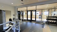 This single-floor fitness center is thoughtfully designed to accommodate a variety of workout styles. The space is divided into distinct zones separated by large, floor-to-ceiling glass panel partitions with sleek black framing, allowing for visual openness while maintaining functional separation between areas. The main workout area features rubberized flooring and houses strength training equipment, including a multi-function cable machine and resistance-based exercise stations. Through the glass partitions, a dedicated studio or stretching room is visible, equipped with stability balls, a wall-mounted ballet barre, and additional fitness accessories — ideal for flexibility, balance, or group exercise sessions. Recessed lighting throughout the ceiling ensures consistent, even illumination across both zones. The neutral color palette of white walls and light-toned finishes contributes to a clean, modern aesthetic. The overall layout is practical and well-organized, providing users with a comfortable and distraction-free environment suitable for both strength training and low-impact exercise activities.