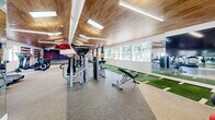 Located at 4 Liberty Lane in South Portland, Maine, this single-floor fitness and recreation facility offers a thoughtfully designed space for athletic training and leisure. The expansive exercise room is the centerpiece, featuring a warm wood-paneled vaulted ceiling with skylights and linear lighting that fills the area with a bright, energetic ambiance. Durable speckled rubber flooring runs throughout the main training zone, where a comprehensive array of Life Fitness strength machines, cable equipment, and cardio machines — including ellipticals and stationary bikes — are arranged for optimal use. A dedicated artificial turf lane, marked with field-style yardage lines, provides a functional surface for agility drills and sled training. At one end of the facility, a full-size boxing ring commands attention, surrounded by bold red pendant lighting, wall-mounted dumbbells, and a flat-screen TV, giving this zone an arena-like feel. Vintage sports posters and bold geometric accent walls add character throughout. A separate lounge area transitions to herringbone hardwood flooring and features a billiards table, a fireplace with textured blue accent wall, and ample natural light from large windows with classic plantation shutters — offering a refined space to unwind after a workout.