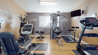 This single-level fitness center is designed to support a well-rounded exercise routine in a compact, functional layout. The space is divided into two connected zones: a main workout area and a transitional entry corridor. The main exercise area features wood-look flooring throughout, with rubber mats strategically placed beneath each piece of equipment for stability and floor protection. The walls are painted in a two-tone scheme — warm tan and charcoal gray — and are adorned with bold motivational typography, including words such as "Passion," "Strength," "Core," "Breathe," "Happy," and "Life," creating an energizing atmosphere. Equipment includes a treadmill, elliptical trainer, stationary bike, multi-function cable machine, flat bench, and a knee raise/dip station, offering options for both cardiovascular and strength training. A wall-mounted flat-screen television provides entertainment during workouts. Natural light enters through blinds-covered windows, complemented by overhead fluorescent lighting. A hand sanitizer dispenser and water cooler are conveniently accessible. The entry corridor, with vaulted ceiling accents and a seating chair, provides a transitional area before entering the main exercise floor, contributing to the overall organized and welcoming character of the facility.