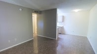 This single-story space presents a straightforward, functional layout with an open living area that flows directly into the kitchen zone. The living area features durable tile-style flooring in a warm neutral tone, complemented by gray-painted walls and white trim. A textured ceiling adds subtle dimension overhead, while a ceiling-mounted HVAC vent ensures climate control throughout the space. The partial wall dividing the living and kitchen areas provides a degree of separation while maintaining an open feel. The kitchen is visible from the main living space and includes light wood cabinetry and a countertop with what appears to be a sink, offering practical functionality in a compact arrangement. A hallway visible from the main room leads to additional areas of the unit, suggesting a practical and efficient floor plan. White walls in the kitchen area contrast with the gray tones of the living space, creating a visual distinction between zones. The overall aesthetic is clean and neutral, providing a blank canvas that is well-suited for personalization.