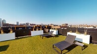 This rooftop amenity level offers a well-appointed outdoor retreat designed for both relaxation and socializing. The expansive open-air terrace is finished with large-format porcelain pavers and features a central fire pit surrounded by modern angular lounge chairs, creating an inviting gathering space. Sleek white rectangular planters filled with ornamental grasses provide both visual interest and subtle boundary definition throughout the deck.

A separate turf lawn area offers a lush green contrast to the hard-surface terrace, furnished with contemporary reclining sun loungers — ideal for daytime relaxation. The rooftop commands sweeping panoramic views of the surrounding urban skyline, with city buildings and open sky visible in every direction, lending the space a distinctly elevated and open ambiance.

Access to the rooftop is provided through a compact interior vestibule, clad in dark corrugated metal siding that gives the structure a sharp, industrial-modern aesthetic. Floor-to-ceiling glass sliding doors connect the interior access room to the terrace, flooding it with natural light and framing striking views of the cityscape beyond. The interior transition space features grey tile flooring, crisp white walls, elevator access, and climate control — providing a functional and clean entry point to the rooftop amenity.