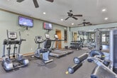 This fitness facility in Columbus, Georgia is a comprehensively outfitted exercise space designed to support a wide range of workout routines. The single-level layout provides generous open floor space covered in durable rubber flooring, ideal for both equipment use and free movement. Soft blue-gray walls are complemented by warm wood trim along the crown molding and door frames, creating a polished yet functional environment.

The cardio section features multiple treadmills and elliptical trainers from True Fitness, as well as a stationary bike and rowing machine, all positioned for easy access. Wall-mounted flat-screen televisions are positioned throughout the room for entertainment during workouts. The strength training area includes a multi-functional cable and power rack system, a dumbbell rack with a full range of weights, and an adjustable bench.

Full-length mirrors line one wall, visually expanding the space and allowing users to monitor their form. A medicine ball rack and stability balls add functional fitness options. French doors along one wall invite natural light and provide outdoor access. Recessed lighting and dual ceiling fans ensure the space remains well-lit and comfortable. A hallway connects to an additional room, adding utility to the overall facility.