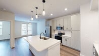 Located at 1811 N Greenville Ave in Richardson, TX, this thoughtfully designed one-bedroom, one-bathroom residence offers a clean, contemporary aesthetic throughout its efficiently utilized single floor. The open-concept kitchen and living area serves as the heart of the home, anchored by a large quartz-topped island with an undermount sink and pendant lighting above. The kitchen is outfitted with gray shaker-style cabinetry, white subway tile backsplash, stainless steel appliances including a French door refrigerator and range with microwave, and a built-in desk nook with wine rack — ideal for working from home. Wood-look flooring flows seamlessly throughout the space, complementing the bright, neutral color palette. The living area benefits from natural light through French doors that open to an outdoor view, with a ceiling fan adding comfort. The bedroom is a serene, well-proportioned retreat featuring a large window, ceiling fan, and wood-look flooring. The bathroom includes a soaking tub with tiled surround, and connects directly to a dedicated walk-in closet with built-in shelving. Clean lines and modern hardware finishes tie the entire space together cohesively.