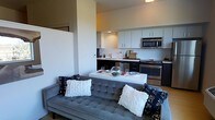 This single-floor residence showcases a thoughtfully designed open-concept layout that connects the living, dining, and kitchen areas into one cohesive and functional space. The living area is anchored by a tufted gray velvet sofa adorned with decorative pillows, creating a comfortable and contemporary seating arrangement. Adjacent to the living space, a compact white dining table provides an intimate setting for meals, flowing naturally into the kitchen. The kitchen is outfitted with sleek dark lower cabinetry contrasted by crisp white upper cabinets, complemented by a classic white subway tile backsplash that adds a clean, timeless aesthetic. Stainless steel appliances, including a built-in microwave and a gas range with oven, lend a modern, polished finish to the culinary space. Recessed lighting illuminates the kitchen, while the overall layout maximizes efficiency without sacrificing style. The neutral color palette of warm whites, grays, and dark accents creates a balanced and inviting ambiance throughout. The open design ensures a seamless visual connection between all areas, making the space feel airy and well-proportioned — ideal for both everyday living and casual entertaining.