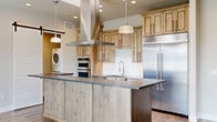 This thoughtfully designed single-floor residence offers a seamless blend of modern style and functional living across a comfortable footprint. Wide-plank hardwood flooring flows throughout, anchoring the open-concept layout and connecting each space cohesively.

The kitchen is a standout centerpiece, showcasing a large island with distressed wood paneling and dark stone countertops, rustic knotty alder cabinetry, stainless steel appliances including a built-in refrigerator and double wall ovens, and a striking ceiling-mounted range hood. Pendant lighting adds warmth above the island, while a subway tile backsplash and a professional-style faucet complete the refined look.

The living area is bright and welcoming, featuring a sleek linear fireplace set into a bold stone-clad wall, a built-in shelving unit with cabinetry, and large windows that invite natural light. A sliding barn door adds a charming architectural detail, while a set of glass patio doors connects the interior to the outdoors.

The bedroom offers a calm retreat with neutral tones and wood flooring. A well-organized walk-in closet with built-in shelving and hanging rods provides generous storage. Both bathrooms are finished with tile flooring, vessel sinks, wood vanities, and framed mirrors. An in-unit laundry area with a stacked washer and dryer adds everyday convenience.