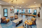 This single-floor community lounge is designed to encourage social interaction through a series of distinct yet open activity zones. The space is anchored by a dramatic glass-panel garage door at the entry, flooding the front seating area with natural light and offering views of the surrounding greenery. A sculptural, multi-arm brass chandelier hangs above this entry zone, where leather armchairs and a concrete coffee table create a relaxed gathering spot.

Moving deeper into the space, a full-length shuffleboard table serves as a central recreational feature, flanked by additional lounge areas with tan leather chairs and blue-toned area rugs that add warmth to the polished tile flooring. Bold, colorful artwork decorates the walls throughout, contributing to the space's energetic aesthetic.

A dedicated lounge corner features teal wingback chairs, a tufted yellow ottoman, and a wall-mounted flat-screen TV, ideal for casual viewing and conversation. Recessed lighting and pendant fixtures maintain consistent, inviting illumination throughout.

The kitchenette area offers white quartz countertops, a stainless steel sink, a wine refrigerator, and a large window overlooking an outdoor patio, while a dining counter with bar seating provides a functional social hub. Abundant windows throughout ensure natural light reaches every corner of this well-appointed communal space.