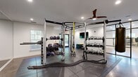 Located at 1030 Jack Primus Rd in Charleston, SC, this single-story fitness facility offers a thoughtfully designed workout environment spread across two dedicated exercise rooms and a connecting hallway. The space is bright and modern, featuring crisp white walls, recessed lighting, and stylish ceiling fans throughout that contribute to a clean, functional ambiance.

The first exercise room is a strength-focused space with dark rubber tile flooring and full-wall mirrors that amplify the sense of openness. It is outfitted with a multi-functional cable and functional trainer rig, battle ropes, TRX suspension systems, a punching bag, medicine balls, and a comprehensive set of kettlebells — all organized on dedicated racks. Natural light filters in through windows, while a wall-mounted flat-screen TV adds a modern touch.

The second exercise room features light hardwood-style flooring and is connected to the strength room via a striking black-framed glass partition wall. This area is equipped with Peloton spin bikes, rowing machines, treadmills, and a dumbbell rack with bench, making it ideal for both cardio and accessory training. A connecting hallway with polished concrete floors ties the two rooms together and provides access to the facility's utilities and entry points.
