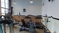 This dedicated fitness space is designed to support a well-rounded exercise routine in a focused, functional environment. The room is outfitted with a selection of SportsArt brand equipment, including treadmills, a stationary bike, and a weight bench, providing options for both cardiovascular training and strength conditioning. A set of free weights adds further versatility to the workout possibilities available. One of the room's most distinctive design features is a bold, black heartbeat line mural that runs along the two-toned walls — the upper half painted in a crisp white and the lower half in a warm tan — giving the space an energetic and purposeful character. Pendant-style light fixtures hang from the ceiling, providing warm, even illumination throughout the room. The carpeted flooring offers a degree of comfort underfoot, while a motivational poster on the wall adds to the gym's encouraging atmosphere. The single-floor layout keeps everything accessible and easy to navigate, making this fitness room a practical and well-appointed space for regular exercise.