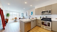 This thoughtfully designed single-floor residence offers a cohesive open-concept layout that seamlessly connects the kitchen, dining, and living areas. The kitchen is a standout feature, equipped with stainless steel appliances including a gas range, built-in microwave, dishwasher, and refrigerator. Flat-front taupe cabinetry pairs elegantly with crisp white countertops, while light hardwood flooring runs throughout the main living spaces, enhancing the bright and airy atmosphere. A peninsula with bar seating provides additional casual dining space alongside a dedicated dining area accommodating a round table with ample seating. The living room is bathed in natural light from a large multi-pane window offering urban views, creating an inviting, comfortable retreat. The bedroom continues the clean aesthetic with soft neutral walls, light hardwood floors, and a generously sized window that draws in natural light and city views. The bathroom features large-format stone-look tile flooring, a gray vanity with modern black fixtures, a wide framed mirror, and a combination tub and shower. A dedicated laundry closet houses a stacked full-size washer and dryer for added convenience. Recessed lighting throughout and a clean, minimalist palette give the entire space a polished, contemporary feel.