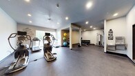This fitness center offers a comprehensive workout environment designed to accommodate a wide range of exercise routines. The space features a textured ceiling with recessed lighting throughout, complemented by large windows along one wall that invite natural light and views of the surrounding greenery, creating an open and energizing atmosphere.

The cardio zone is prominently positioned near the windows and includes treadmills and an elliptical trainer, allowing users to enjoy natural light during their workouts. Toward the center and rear of the space, structural columns subtly divide the floor plan into functional zones without disrupting the overall openness.

A dedicated free weights area houses a full rack of dumbbells alongside a weight bench, catering to strength training needs. Adjacent to this, a cable and resistance machine provides options for a variety of targeted exercises. The exercise and flexibility zone is outfitted with stability balls in multiple sizes, offering options for core training and stretching routines.

The flooring throughout is a durable, dark rubberized material suited for high-traffic fitness use, while the neutral-toned walls keep the environment clean and focused. Motivational wall art adds a welcoming touch. Overall, the space is thoughtfully organized to support both cardio and strength training in a bright, functional setting.