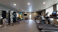 This fitness center offers a thorough and thoughtfully arranged workout environment spread across a single floor. The space is finished with durable rubber-speckled flooring suited for high-traffic exercise use, smooth grey walls, and recessed lighting that keeps the room bright and functional throughout.

The cardio section is prominently featured along the windowed wall, where multiple treadmills and elliptical trainers equipped with individual digital display screens are lined up to provide an engaging workout experience. Natural light filters in through large windows fitted with roller shades, offering views to the exterior while maintaining a comfortable atmosphere. A rowing machine and stationary bike round out the cardio offerings.

On the strength training side, the facility is well-stocked with a multi-function cable machine, a full rack of dumbbells spanning a wide range of weights, kettlebells in various sizes, and an assortment of stability and medicine balls. Adjustable workout benches are positioned throughout the open floor plan for versatile use.

Wall-mounted flat-screen televisions are placed at strategic points around the room for entertainment during workouts. A split-system air conditioning unit ensures climate control year-round. The overall design is clean, modern, and purposeful, providing users with a complete fitness experience in a comfortable, well-maintained setting.