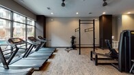 This modern fitness center is designed to support a full range of workout routines in a sleek, well-appointed environment. The space is finished with neutral-toned carpeting, recessed lighting, and ceiling fans throughout, creating a comfortable and functional atmosphere for exercise.

The cardio zone features a row of treadmills positioned alongside floor-to-ceiling windows that offer views of the surrounding outdoor courtyard, flooding the area with natural light. Nearby, elliptical machines, stationary bikes, and rowing machines provide a comprehensive selection of low-impact cardio options.

The strength training area is generously outfitted with multiple selectorized cable and resistance machines, catering to a variety of muscle groups. A dedicated free weights section includes a full rack of kettlebells and dumbbells, accompanied by benches for seated and supine exercises. A multi-functional power rack with pull-up bars and cable attachments anchors the functional training zone, ideal for compound and bodyweight movements.

A connecting corridor lined with large mirrors leads to additional areas of the facility, with dark-toned walls and accents lending a contemporary, upscale aesthetic throughout. Exposed dark beam details and reflective mirror panels enhance the sense of depth and space, making this fitness center a standout amenity.