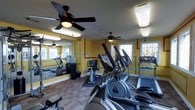 This fitness center offers a practical and energetic environment for exercise. The room is finished with bold yellow walls accented by colorful geometric borders in teal and red, creating a lively atmosphere. Multiple ceiling fans are installed throughout, promoting air circulation and comfort during workouts. Large windows along one wall allow natural light to fill the space, contributing to an open and inviting feel. A full-length mirror runs along one side of the room, visually expanding the area and allowing users to monitor their form. The flooring features a combination of carpet and rubber mat sections, providing both comfort and stability underfoot. The cardio equipment selection is comprehensive, including elliptical trainers, treadmills, and a stationary bike, all arranged to allow ease of movement between machines. Overhead fluorescent lighting supplements the natural light, ensuring the space remains well-lit at all hours. The single-floor layout keeps everything accessible and straightforward, making it a functional and well-appointed fitness facility suited for a range of cardio-focused workouts.