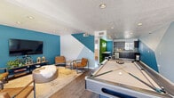 Located at 919 East Lemon Street in Tempe, Arizona, this single-floor community space offers a well-designed and energetic environment across three distinct areas. The living and game room serves as the centerpiece, featuring bold geometric wall murals in teal and white that create a striking visual statement. A full-size pool table anchors the recreational side of the room, while a seating area with leather accent chairs, a wall-mounted flat-screen TV, and a lush vertical moss wall panel add personality and comfort. Warm hardwood-style flooring runs throughout, tying the space together seamlessly. A high-top bar table with stools provides an additional social hub near the kitchen transition. The kitchenette is outfitted with dark, modern cabinetry, a mosaic tile backsplash, a stainless steel sink, a compact refrigerator, and a coffee station — ideal for casual refreshment and gathering. Natural light filters in through a window overlooking the streetscape. The dedicated office area is clean and functional, furnished with a long workstation, multiple computer terminals, a printer, and ergonomic chairs. A bold branded accent wall in teal with a gold logo reinforces the space's identity. Recessed lighting throughout ensures a bright, welcoming atmosphere in every area.