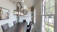 This single-floor space exudes a sophisticated and inviting ambiance, thoughtfully designed with a blend of classic and contemporary elements. The dining area serves as a focal point, anchored by a dark-stained rectangular dining table surrounded by upholstered charcoal chairs that add both comfort and style. Above, a striking geometric pendant light fixture in a dark metal finish commands attention, casting a warm glow over the space.

A beautifully crafted white fireplace mantel serves as an elegant centerpiece along the main wall, flanked by symmetrical built-in shelving units that offer tasteful display space for decorative accents, including ceramic vases and curated décor pieces. A round decorative mirror with a textured frame is mounted above the mantel, adding depth and visual interest to the composition.

Natural light pours generously into the room through large multi-pane windows, creating a bright and airy atmosphere while offering views to the exterior. Neutral warm-toned walls, crisp crown molding, and a patterned area rug beneath the dining set tie the design together seamlessly. The overall aesthetic is polished and cohesive, making this space well-suited for both everyday gatherings and formal entertaining.