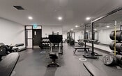 Located at 10023 104 Street NW in downtown Edmonton, Alberta, this fitness facility offers a generously sized, single-floor exercise room designed to support a full range of workout routines. The space features a clean, modern aesthetic with crisp white walls, recessed lighting, and a combination of dark rubber and polished flooring that gives the gym a sleek, professional appearance.

The cardio section is well-appointed with treadmills, elliptical machines, stationary bikes, and a rowing machine, providing users with a variety of low- and high-impact options. The strength training area is equally comprehensive, featuring multiple power racks loaded with Olympic barbells and weight plates, adjustable benches, and a multi-station cable machine suited for a wide range of functional exercises. A dedicated dumbbell rack rounds out the free weights section.

Wall-to-wall mirrors line portions of the facility, enhancing the sense of openness and allowing users to monitor their form during training. The layout is thoughtfully organized to separate cardio and strength zones, ensuring a smooth and efficient flow throughout the space. Ceiling-mounted HVAC units maintain climate control, while recessed pot lights provide bright, even illumination. A glass-panel door allows natural light to filter into the cardio area, adding a welcoming touch to this fully equipped fitness space.