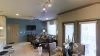 This single-story space showcases an open-concept layout that seamlessly connects the living and dining areas. The living room is anchored by a bold teal-blue accent wall that adds a pop of color against the neutral tones throughout the space. Track lighting installed on the ceiling provides adjustable illumination, complemented by a ceiling fan with integrated lighting for added comfort and airflow. A wall-mounted flat-screen television is positioned above a dark wood media console, while a plush sofa with decorative pillows and a matching armchair create a welcoming seating arrangement around a wooden coffee table. A patterned area rug defines the living space beneath.

The dining area, partially visible to the right, features a well-set table adorned with decorative floral centerpieces, place settings, and elegant tableware. Natural light filters through multiple windows dressed with layered curtains and blinds, including a larger sliding or paneled door that brings in additional brightness. The cream-colored carpeting throughout adds warmth, and the overall palette of browns, blues, and neutrals creates a cohesive and inviting atmosphere suited for both everyday living and entertaining.