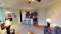 This single-floor residence showcases an open-concept design that effortlessly connects the living area and kitchen, creating a spacious and functional flow throughout. The living room features plush carpeting and is anchored by a ceiling fan, adding both comfort and style to the space. Crown molding runs along the ceiling, lending a refined architectural touch to the interior.

The kitchen is a standout feature, highlighted by rich dark cabinetry that extends to the upper walls, complemented by a suite of black appliances including a refrigerator, microwave, and range. A kitchen island with a light-colored countertop serves as both a prep space and a casual dining area, accompanied by two bar-height stools. Pendant lighting hangs above the island, while track lighting illuminates the cabinetry and countertops below.

The transition between the carpeted living area and the hardwood-style flooring of the kitchen creates a natural visual boundary between the two zones. Neutral wall tones throughout the space enhance the sense of openness and allow the darker cabinetry and warm wood flooring to stand out. The overall ambiance is warm, cohesive, and well-suited for both everyday living and casual entertaining.