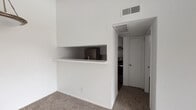 This single-story space is designed with a practical, open layout that connects the living area, kitchen, and bedroom in a straightforward flow. The living room is carpeted in a neutral, textured carpet and is illuminated by natural light entering through a sliding glass door fitted with vertical blinds, which provides direct access to an outdoor patio area. The open sightlines and light-colored walls contribute to an airy, clean atmosphere throughout.

The kitchen is accessible via a pass-through window with a built-in counter ledge, creating a visual and functional connection to the living space. Inside the kitchen, white cabinetry lines both walls, complemented by light countertops, a stainless steel refrigerator, a dishwasher, and a sink — all set above dark hardwood-style flooring that contrasts with the rest of the unit.

A dining area adjacent to the living space features a hanging pendant light fixture with a brushed metal finish, adding a subtle decorative element to the otherwise understated interior.

The bedroom is carpeted and includes a mirrored sliding closet, maximizing the sense of space. Textured ceilings are present throughout, and ceiling-mounted HVAC vents provide climate control. The overall palette is neutral and clean, offering a blank canvas well-suited for personalization.