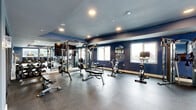 Located at 20 Grand Ave in Englewood, NJ, this well-appointed fitness center offers a comprehensive workout facility spread across a single floor. The space is thoughtfully divided into distinct zones designed to support a wide range of exercise routines.

The main exercise room is the centerpiece of the facility, finished with durable rubber flooring and bold navy blue walls that lend an energetic atmosphere. Large windows allow natural light to fill the space while offering views of the surrounding area. The room is equipped with a robust selection of strength and cardio equipment, including cable machines, weight benches, spin bikes, and a treadmill. Full-length mirrors line the walls to maximize the sense of space and support proper form during workouts. A built-in water fountain and a wall-mounted flat-screen TV add convenience and comfort.

Adjacent to the main gym is a bright, multipurpose studio room featuring light hardwood flooring, crisp white walls, and large mirrors. This area is well-suited for yoga, stretching, or light functional training, and includes stability balls and resistance equipment. Fitness-themed wall art adds a motivating aesthetic touch.

The connecting hallways link the rooms cleanly and efficiently, contributing to a smooth flow throughout the facility. Together, the areas create a complete, well-rounded fitness environment suitable for a variety of training needs.