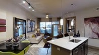 This thoughtfully designed single-floor residence showcases a contemporary open-concept layout that connects the kitchen, dining, and living areas into one cohesive, flowing space. The kitchen takes center stage with a sleek island topped in crisp white quartz, a stainless steel undermount sink, and elegant pendant lighting that draws the eye upward. Dark cabinetry provides a striking contrast to the bright countertops, lending the kitchen a modern, sophisticated character.

The living area is generously proportioned, anchored by a plush neutral sofa accented with vibrant throw pillows and complemented by a patterned area rug. A dramatic arc floor lamp and recessed track lighting work in tandem to create a warm, layered ambiance throughout the space. Large windows dressed with floor-length drapes flood the interior with natural light while offering views to the outside.

Adjacent to the living room, a glimpse into the bedroom reveals a ceiling fan and refined furnishings, maintaining the cohesive design language found throughout. Hardwood-style flooring runs seamlessly across the space, enhancing its open, airy feel. The overall ambiance is modern and inviting, blending functional design with carefully curated aesthetic details that make this residence both practical and visually compelling.