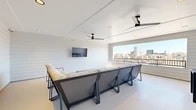 This rooftop lounge is a thoughtfully designed semi-enclosed retreat that offers an elevated urban experience. The space is defined by crisp white shiplap walls that lend a clean, modern coastal aesthetic, complemented by a smooth white ceiling outfitted with dual black ceiling fans and recessed lighting to ensure comfortable airflow and ambiance throughout.

A large open-frame window dominates one wall, framing sweeping views of the surrounding city skyline and flooding the interior with natural light. The polished concrete-style flooring grounds the space with a sleek, durable finish suited for high-traffic communal use.

The lounge is organized into distinct seating zones. A generous L-shaped sectional sofa with plush neutral cushions anchors the primary lounge area, centered around a low-profile coffee table atop a geometric area rug — ideal for casual conversation or entertainment. A wall-mounted flat-screen TV serves as a focal point opposite the seating. Toward the perimeter, high-top bistro tables paired with woven bar chairs provide an alternative setting for more upright socializing or dining.

A vibrant botanical wall art piece introduces a pop of color, adding personality to the otherwise neutral palette. A yellow accent door provides a bold architectural contrast at the entry point. Overall, the space presents a well-appointed, versatile lounge environment with strong urban views and a welcoming modern atmosphere.