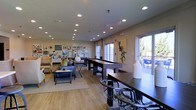This single-floor community space is designed to foster social interaction and relaxation in a well-appointed, open-plan layout. The interior is anchored by a full-length shuffleboard table, serving as a central recreational focal point that draws residents together. Adjacent to the game area, a comfortable lounge section features plush seating, including armchairs and a sofa arranged around a coffee table atop a neutral area rug, creating an inviting space for casual conversation or unwinding.

The room is finished with warm-toned hardwood flooring that runs throughout, complementing the light beige walls and contributing to a bright, airy atmosphere. Recessed lighting is evenly distributed across the ceiling, ensuring the space is well-lit at all times. A curated gallery wall displaying framed artwork and decorative signage adds personality and visual interest to one end of the room.

Large windows along the perimeter flood the interior with natural light and offer views of an outdoor seating area, where patio furniture is visible, hinting at additional amenity space beyond. A communal high-top table with stylish bar-stool seating provides an alternative spot for gathering or casual dining. Overall, the space balances recreational functionality with a comfortable, welcoming ambiance suited for a variety of social activities.