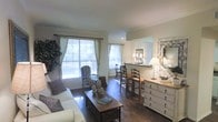 This single-floor space showcases a well-appointed interior defined by rich, dark hardwood flooring that extends throughout the open-concept layout. The living area is anchored by a comfortable sofa adorned with patterned accent pillows, complemented by a classic wooden coffee table and an elegantly upholstered wingback chair. Two generously sized windows dressed with sheer curtains flood the room with abundant natural light, creating a bright and inviting atmosphere.

A partial wall with a pass-through opening connects the living area to an adjacent dining space, where a wooden table and chairs are positioned beneath warm lamp lighting. This architectural detail adds depth to the layout while maintaining a sense of openness between the two areas.

A large white dresser with detailed hardware lines one wall, topped with decorative mirrors and table lamps that add warmth and visual interest. Crown molding along the ceiling contributes a refined, finished look to the space. The overall ambiance is warm and traditionally styled, combining functional furniture arrangements with classic decorative touches to create a cohesive and comfortable environment.