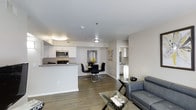 This single-floor residence offers a thoughtfully designed open-concept layout that seamlessly connects the kitchen, dining, and living areas, creating a spacious and functional living environment. The kitchen is outfitted with stainless steel appliances, including a microwave, range, and refrigerator, complemented by clean white cabinetry and a peninsula counter that serves as both a workspace and a casual divider between the kitchen and the rest of the living space. The flooring throughout features rich, dark hardwood-style planks that provide a warm contrast to the light gray and white walls, lending the space a modern and cohesive aesthetic. The dining area accommodates a four-seat table set in dark tones, positioned beneath a ceiling-mounted light fixture that adds a subtle focal point to the space. The living area is furnished with a sleek gray sofa and a glass-top coffee table, offering a comfortable and contemporary retreat. Recessed and flush-mount lighting throughout ensures the space remains bright and inviting. The open layout and neutral color palette create a versatile environment that feels airy and well-proportioned, making it ideal for both everyday living and entertaining.