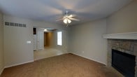 This two-story space offers a straightforward and functional design with a neutral color palette throughout. The main living area is carpeted and generously sized, providing a comfortable and versatile space for everyday use. A ceiling fan with an integrated light fixture anchors the room, ensuring both airflow and ambient lighting. A fireplace mantel is visible along one wall, adding a classic focal point to the living area. The room opens through an arched passageway into an adjacent space, which features a pendant light and connects to additional rooms beyond. A door visible in the background leads to further areas of the home, hinting at a well-connected interior layout. Natural light filters in through a window, brightening the neutral walls and light-toned ceiling. Tile flooring transitions from the carpet near the fireplace area, suggesting a defined entryway or transition zone. The overall ambiance is calm and understated, with clean lines and simple finishes that provide a versatile backdrop. Spanning two floors, this space offers room for varied uses and comfortable day-to-day living.