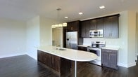 This single-floor space showcases a thoughtfully designed, open-concept layout centered around a stunning kitchen. The kitchen is a standout feature, combining rich dark espresso cabinetry that extends to the ceiling with crisp white countertops for a striking contrast. A generously sized curved island anchors the space, complete with an integrated sink and pendant lighting overhead — a sleek, multi-bulb chrome fixture that adds a contemporary touch.

The kitchen is fully equipped with stainless steel appliances, including a French door refrigerator, an electric range, and a built-in microwave, all seamlessly integrated into the cabinetry for a cohesive, polished look. Recessed lighting throughout the ceiling ensures the space is well-lit, complementing the modern aesthetic.

The rich, dark hardwood flooring runs throughout, grounding the space and adding warmth against the lighter walls and countertops. The open layout connects the kitchen to the adjacent living areas, creating a fluid and functional flow. Clean lines, quality finishes, and a neutral color palette come together to give this space a modern, refined ambiance that is both practical and visually impressive.
