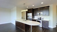 This single-floor space showcases a thoughtfully designed, open-concept layout centered around a stunning kitchen. The kitchen is a standout feature, combining rich dark espresso cabinetry that extends to the ceiling with crisp white countertops for a striking contrast. A generously sized curved island anchors the space, complete with an integrated sink and pendant lighting overhead — a sleek, multi-bulb chrome fixture that adds a contemporary touch.

The kitchen is fully equipped with stainless steel appliances, including a French door refrigerator, an electric range, and a built-in microwave, all seamlessly integrated into the cabinetry for a cohesive, polished look. Recessed lighting throughout the ceiling ensures the space is well-lit, complementing the modern aesthetic.

The rich, dark hardwood flooring runs throughout, grounding the space and adding warmth against the lighter walls and countertops. The open layout connects the kitchen to the adjacent living areas, creating a fluid and functional flow. Clean lines, quality finishes, and a neutral color palette come together to give this space a modern, refined ambiance that is both practical and visually impressive.