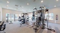 Located in Humble, TX, this single-floor fitness center is designed to support a full range of workout routines in a bright, welcoming environment. The main workout area is generously sized, housing an extensive array of strength-training machines, including cable systems, multi-functional weight stations, and benches, all arranged across a durable carpet-and-hardwood floor combination. Ceiling fans and recessed lighting keep the space comfortable and well-lit, while large windows along multiple walls flood the room with natural light and offer views of the surrounding outdoor area, including a pool courtyard. A dedicated row of True elliptical machines and additional cardio equipment lines the window-facing wall, making workouts both functional and scenic. A dual-station water fountain with a bottle-filling option is conveniently integrated into the space, set against a clean white brick accent wall. A separate yoga and studio room features warm hardwood flooring, a tray ceiling with a ceiling fan, recessed lighting, floor-to-ceiling mirrors, a large mounted flat-screen TV, rowing machines, and yoga mats — creating an ideal setting for group classes or individual training. The overall ambiance is clean, modern, and purposefully designed to encourage an active lifestyle.