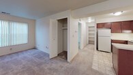 This single-floor residence offers a practical and well-organized layout designed for comfortable everyday living. The kitchen is a standout feature, showcasing rich dark mahogany cabinetry that extends to the ceiling, providing ample storage throughout. It is equipped with a full suite of white appliances, including a refrigerator, electric range, and dishwasher, all complemented by light-toned countertops and tile flooring that add brightness to the space. A peninsula counter creates a natural transition into the adjacent living and dining area, which is finished with wall-to-wall carpeting for a warm, comfortable feel. The spacious living area features a front entry door and ceiling-mounted lighting, creating a clean, open environment with room for flexible furniture arrangements. A dedicated bedroom area offers generous proportions, vertical blind window treatments for light control, and access to a closet with built-in shelving. A hallway connects the main living areas and features additional built-in shelving, ideal for organized storage. The overall design is neutral and move-in ready, with consistent light-toned walls throughout that contribute to a clean, airy ambiance across the entire floor plan.