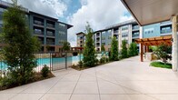 Located at 3640 Mapleshade Ln in Plano, Texas, this ground-level outdoor amenity and commercial space is set within a contemporary multi-story mixed-use complex. The centerpiece of the outdoor area is a resort-style swimming pool, enclosed by an iron fence and surrounded by manicured landscaping, including tall evergreen trees and neatly trimmed shrubs that provide both privacy and greenery. Poolside seating with colorful lounge chairs and shaded umbrellas contributes to a welcoming, resort-inspired atmosphere. A spacious concrete courtyard extends from the pool area, offering ample open space for recreation and leisure. A putting green is also visible within the courtyard, adding to the variety of outdoor amenities available. The multi-story residential buildings surrounding the courtyard feature a modern architectural design, combining dark paneling, brick accents, and large windows with private balconies that overlook the amenity space below. The ground-floor commercial frontage is clad in natural limestone masonry, with floor-to-ceiling glass storefronts that allow views into the interior and out toward the pool courtyard. This combination of thoughtful landscaping, modern architecture, and diverse amenities creates a vibrant, community-oriented environment in the heart of Plano.