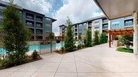 Located at 3640 Mapleshade Ln in Plano, Texas, this ground-level outdoor amenity and commercial space is set within a contemporary multi-story mixed-use complex. The centerpiece of the outdoor area is a resort-style swimming pool, enclosed by an iron fence and surrounded by manicured landscaping, including tall evergreen trees and neatly trimmed shrubs that provide both privacy and greenery. Poolside seating with colorful lounge chairs and shaded umbrellas contributes to a welcoming, resort-inspired atmosphere. A spacious concrete courtyard extends from the pool area, offering ample open space for recreation and leisure. A putting green is also visible within the courtyard, adding to the variety of outdoor amenities available. The multi-story residential buildings surrounding the courtyard feature a modern architectural design, combining dark paneling, brick accents, and large windows with private balconies that overlook the amenity space below. The ground-floor commercial frontage is clad in natural limestone masonry, with floor-to-ceiling glass storefronts that allow views into the interior and out toward the pool courtyard. This combination of thoughtful landscaping, modern architecture, and diverse amenities creates a vibrant, community-oriented environment in the heart of Plano.