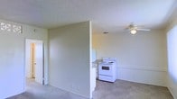 This single-floor space features an open-concept layout that connects the living area to the kitchen, creating a functional and straightforward floor plan. The living area is finished with neutral carpet flooring and light-toned walls, contributing to a clean, understated aesthetic. A ceiling fan is centrally positioned overhead, providing air circulation throughout the shared space.

The kitchen is partially separated by a half-wall partition, offering a degree of definition between the cooking and living zones while maintaining an open feel. It is equipped with a white electric range and includes counter space and cabinetry in a simple, utilitarian arrangement. Light tile flooring in the kitchen complements the neutral palette carried throughout the unit.

Textured popcorn ceilings are present throughout, consistent with standard residential construction of its era. The overall ambiance is minimal and functional, with neutral colors and basic finishes providing a blank canvas. A doorway visible on the left side of the living area suggests access to additional rooms within the unit. The space is well-suited for those seeking a straightforward, low-maintenance living environment.