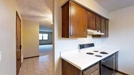 This single-floor space offers a practical and straightforward layout designed for comfortable everyday living. The kitchen is equipped with dark wood upper cabinetry featuring brass-toned hardware, providing ample storage space. White laminate countertops offer a clean contrast against the rich cabinetry tones, while a white electric coil range with a built-in overhead exhaust fan serves as the primary cooking station. The kitchen floor is finished with neutral tile, adding durability to the space. An open passageway connects the kitchen directly to the adjacent living area, creating a sense of flow between the two spaces. The living area is finished with wall-to-wall carpeting in a neutral tone, lending a softer, more comfortable feel underfoot. A window in the living area allows natural light to filter in, brightening the room. Both spaces feature textured ceilings, consistent with the overall functional aesthetic of the unit. The neutral wall tones throughout create a clean, versatile backdrop well-suited to a variety of furnishing styles.