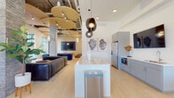 This contemporary single-floor amenity space is thoughtfully designed to blend comfort, style, and functionality in an open, loft-inspired layout. The interior boasts soaring ceilings with exposed industrial ductwork juxtaposed against striking sculptural wooden ceiling medallions, creating a dynamic visual contrast throughout the space.

The lounge area is anchored by a large leather sofa and a sleek linear gas fireplace set against a floor-to-ceiling stone accent wall, with a pair of modern armchairs completing an intimate seating arrangement. Large floor-to-ceiling glass doors and windows flood the area with natural light and provide direct access to an outdoor terrace.

A separate media lounge features bold blue velvet channel-tufted seating arranged around a wall-mounted flat-screen TV, complemented by cascading globe pendant lighting and light wood flooring that runs throughout the space.

The kitchenette is well-appointed with grey cabinetry, a stainless steel refrigerator, a built-in microwave, a sink, and a central island with pendant lighting overhead, making it well-suited for entertaining.

A games area rounds out the space, featuring a full-size billiards table and additional seating, with expansive windows offering views to an adjacent outdoor courtyard. The overall ambiance is sophisticated yet inviting, combining industrial and modern design elements seamlessly.
