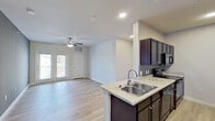 This single-floor residence offers a thoughtfully designed open-concept layout that seamlessly connects the living area and kitchen. The living room features light wood-look flooring, a ceiling fan, recessed lighting, and access to a private exterior door, creating a bright and welcoming atmosphere. The kitchen is designed in a galley style with rich dark espresso cabinetry, light granite-style countertops, a double stainless steel sink, and a full suite of black appliances including a refrigerator, electric range, built-in microwave, and dishwasher. Ample upper and lower cabinetry provides generous storage throughout. A hallway with bold gray accent walls connects the living spaces to the private areas of the home. The bedroom is generously sized, featuring plush carpeting, a ceiling fan, and multiple windows that allow natural light to fill the room. An additional room with gray walls and wood-look flooring offers flexible use as a secondary bedroom or office. The bathroom is finished with a tiled tub-and-shower surround in a slate-toned tile, light wood-look flooring, a dark vanity with granite-style countertop, and chrome fixtures. Clean white walls and consistent flooring choices throughout give the space a cohesive, modern feel.