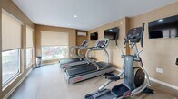 Located at 400 Hoover Way in Woodbridge, NJ, this fitness center offers a well-appointed exercise space designed to support an active lifestyle. The room features light-toned textured walls and large windows fitted with roller shades, allowing natural light to fill the space while maintaining privacy. Light-colored tile flooring runs throughout, contributing to a clean and polished aesthetic.

The fitness center is equipped with multiple Star Trac treadmills and an elliptical machine positioned along the windowed wall, allowing users to enjoy outdoor views during workouts. A stationary spin bike rounds out the cardio offerings. Two wall-mounted flat-screen televisions are strategically placed for visibility from the equipment, enhancing the workout experience.

A wall-mounted air conditioning unit ensures climate control year-round. Sanitation stations, including a hand sanitizer dispenser and a waste receptacle, are conveniently positioned near the entrance, reflecting a commitment to cleanliness. Motivational fitness photography adorns the walls, adding energy to the environment.

The space is accessed through a glass-panel door, with an additional interior door providing secondary access. Recessed ceiling lighting supplements the natural light, ensuring the room remains bright and functional at all hours. Overall, the fitness center presents a tidy, practical, and inviting environment for residents seeking convenient on-site exercise facilities.