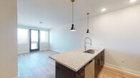 This single-floor unit showcases a thoughtfully designed open-concept layout that seamlessly connects the kitchen and living areas. The kitchen serves as the centerpiece, anchored by a substantial island finished with speckled granite countertops and a navy blue base. A commercial-style pull-down faucet and undermount sink add a professional touch, while two sleek pendant lights with brass accents hang above the island, providing both task lighting and visual interest. The kitchen wall features white upper cabinetry, a subway tile backsplash, a gas range, stainless steel microwave, and a French door refrigerator, all complemented by dark lower cabinetry for a two-tone aesthetic.

Light wood-look flooring flows consistently throughout the space, contributing to an airy and cohesive feel. The open living area offers generous room for furnishings, with large windows allowing natural light to fill the space. A bedroom features a ceiling fan, wide windows with blinds, and the same warm flooring. The bathroom mirrors the unit's design language, presenting a granite vanity countertop, dark-framed mirror, and dark wood-grain cabinetry with matte black hardware.