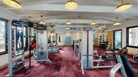 Located in Burlington, North Carolina, this single-floor fitness center offers a thoughtfully designed workout environment that balances functionality with refined aesthetics. The space is finished with rich burgundy carpet tiles throughout, lending a distinctive, upscale character rarely found in community fitness facilities.

The ceiling features exposed beam detailing with a vaulted profile in select areas, adding architectural interest overhead. A series of gold pendant lights hang at varying heights across the space, providing warm, even illumination and contributing to the room's polished atmosphere. Multiple flat-panel televisions are mounted on the walls, offering entertainment during workouts.

Natural light enters generously through several dark-trimmed windows positioned along the walls, offering views of the surrounding community exterior. The open floor plan is organized into distinct functional zones. A dedicated free weight area includes a full rack of dumbbells and multiple barbell stations with weight plates. Strength training is well supported by Life Fitness cable machines, including a Synrgy360 functional trainer. The cardio zone features treadmills, stair climbers, stationary cycles, and rowing machines. Wall-mounted mirrors expand the sense of space and allow users to monitor form. The layout allows ample circulation between equipment, creating a comfortable and efficient workout environment.