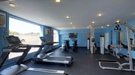 This fitness center offers a clean, functional workout environment designed to accommodate a variety of exercise routines. The space is defined by its striking blue accent walls, which contrast with the crisp white ceiling outfitted with recessed lighting that ensures consistent, even illumination throughout. Large windows along one wall invite natural light and provide views to the outdoors, enhancing the overall energy of the room.

The cardio area is well-appointed, featuring treadmills and a row of stationary cycling bikes, giving users ample options for aerobic training. A wall-mounted flat-screen television is positioned for visibility across the floor, adding an entertainment element during workouts. Full-length mirrors line one wall, contributing both a sense of expanded space and practical utility for form-checking during exercise.

The flooring consists of durable dark rubber matting, a practical and safety-conscious choice suited for heavy equipment and high-traffic use. Sleek stainless steel waste receptacles are neatly placed throughout the room, reflecting an attention to cleanliness and organization. A wall-mounted air conditioning unit ensures climate control for user comfort. Overall, the space presents a tidy, purposeful, and inviting fitness environment suitable for daily use.