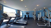 This fitness center offers a clean, functional workout environment designed to accommodate a variety of exercise routines. The space is defined by its striking blue accent walls, which contrast with the crisp white ceiling outfitted with recessed lighting that ensures consistent, even illumination throughout. Large windows along one wall invite natural light and provide views to the outdoors, enhancing the overall energy of the room.

The cardio area is well-appointed, featuring treadmills and a row of stationary cycling bikes, giving users ample options for aerobic training. A wall-mounted flat-screen television is positioned for visibility across the floor, adding an entertainment element during workouts. Full-length mirrors line one wall, contributing both a sense of expanded space and practical utility for form-checking during exercise.

The flooring consists of durable dark rubber matting, a practical and safety-conscious choice suited for heavy equipment and high-traffic use. Sleek stainless steel waste receptacles are neatly placed throughout the room, reflecting an attention to cleanliness and organization. A wall-mounted air conditioning unit ensures climate control for user comfort. Overall, the space presents a tidy, purposeful, and inviting fitness environment suitable for daily use.