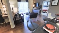 This well-appointed single-floor residence showcases a thoughtfully designed open-concept layout that seamlessly connects the living and dining areas, creating a fluid and inviting atmosphere. Rich, dark hardwood flooring runs throughout, anchoring the space with warmth and sophistication.

The living area is elegantly furnished with sleek, modern seating — including chrome-framed leather chairs — centered around a contemporary coffee table. A media console with built-in cabinetry provides both storage and a focal point for the entertainment setup. Large windows dressed with floor-length curtains allow generous natural light to pour in, brightening the interior and offering a connection to the outdoors.

Accent lighting from stylish table lamps adds warmth during evening hours, complementing the overall modern aesthetic. The neutral wall tones are enhanced by carefully chosen dark furnishings and décor, creating a balanced, contemporary look throughout.

A glimpse into the adjacent bedroom reveals a well-lit, comfortably appointed sleeping space with coordinated furnishings, maintaining the same polished design language found in the main living area. The overall ambiance of the residence is modern, refined, and comfortable — ideal for both everyday living and entertaining.