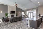 This single-floor communal space is thoughtfully designed with an open, flowing layout that seamlessly connects multiple functional zones. The living area serves as the heart of the space, anchored by a striking floor-to-ceiling stacked-stone fireplace with a built-in TV mount above, flanked by lush greenery and comfortable leather seating arranged on wood-look tile flooring. Vaulted ceilings with recessed lighting and a ceiling fan add volume and airiness to the room, while neutral taupe walls contribute to a warm, welcoming ambiance.

Large windows and glass-panel exterior doors flood the interior with natural light, offering views of the surrounding exterior. A dedicated dining area with a long dark-wood table and chairs sits adjacent to the living space, making the layout well-suited for gatherings.

The kitchenette area features dark espresso cabinetry with sleek hardware, light granite countertops, a white subway tile backsplash, stainless steel appliances including a full-size refrigerator and microwave, and ample storage. A large decorative wall clock adds a rustic accent to the kitchen corridor. A counter-height bar with stools provides additional seating. Together, these elements create a polished, contemporary communal environment ideal for socializing and events.