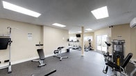 This fitness center is designed to provide a comprehensive workout environment within a single, open floor layout. The space features a drop-panel ceiling with recessed flat-panel lighting that ensures consistent, even illumination throughout. Neutral beige walls contribute to a clean, welcoming atmosphere, while durable rubber-mat flooring offers both safety and comfort underfoot.

The cardio section is equipped with multiple treadmills and an elliptical trainer, all neatly arranged to allow ample space between machines. A wall-mounted flat-screen television is positioned to keep users entertained during workouts. The strength and conditioning area includes adjustable weight benches and free weight equipment, offering a variety of training options. A sliding glass door at the far end of the room allows natural light to fill the space and provides access to an outdoor area, further enhancing the open and airy feel of the facility.

The layout is thoughtfully organized to accommodate simultaneous use by multiple individuals without feeling crowded. A central structural column is seamlessly integrated into the floor plan, and informational signage is posted near the entrance. Overall, this fitness center delivers a functional, well-maintained environment suited for a full range of exercise routines.
