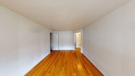 This single-floor unit is characterized by warm-toned hardwood flooring that runs consistently throughout the living spaces and bedrooms, lending a cohesive and classic feel to the interior. The walls are finished in crisp white throughout, creating a clean, neutral backdrop that maximizes the sense of light and openness in each room.

The main living area is generously proportioned, with smooth white ceilings and an open feel. It connects to additional rooms via interior doorways, allowing for a straightforward, functional layout as shown in the overhead floor plan view. Natural light enters through multiple windows fitted with roller shades and sheer curtains.

The bedrooms continue the hardwood flooring and white wall palette. One bedroom features a built-in closet and an outdoor-facing window with views of surrounding greenery. Another bedroom is equipped with a cast-iron radiator beneath a double window, providing both warmth and natural light.

The kitchen is efficiently designed with white cabinetry extending to the ceiling, offering ample storage. It includes a white electric range, refrigerator, and a single-basin sink, all set against light-toned flooring.

The bathroom features a full tub-shower combination, a white vanity with under-sink storage, a wall-mounted mirror, a radiator, and a window for natural ventilation.